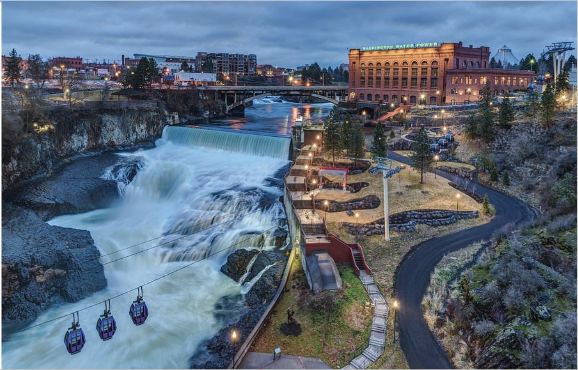 Night view of the Washington Water Power building next to a waterfall with a river, bridge, gondolas, and park area with pathways and lights in Spokane, Washington.