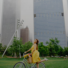 Woman in yellow dress riding a bicycle in a city park with tall buildings and green trees in the background.