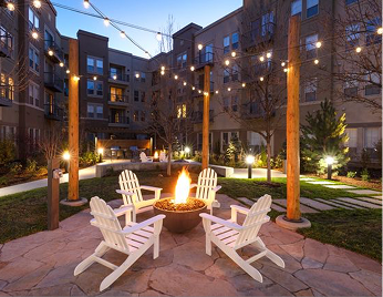 Outdoor courtyard with string lights overhead, a fire pit with flames, four white Adirondack chairs arranged around it, and apartment buildings in the background.