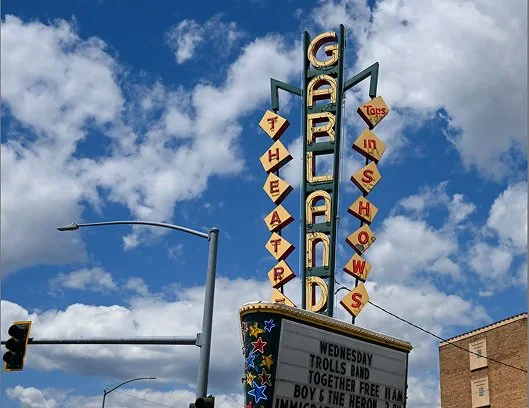 Giant sign with the word "GIGANTIC" vertically in large letters, and "THEATRE" and "SHOWS" displayed in diamond shapes. Blue sky with scattered clouds in the background.