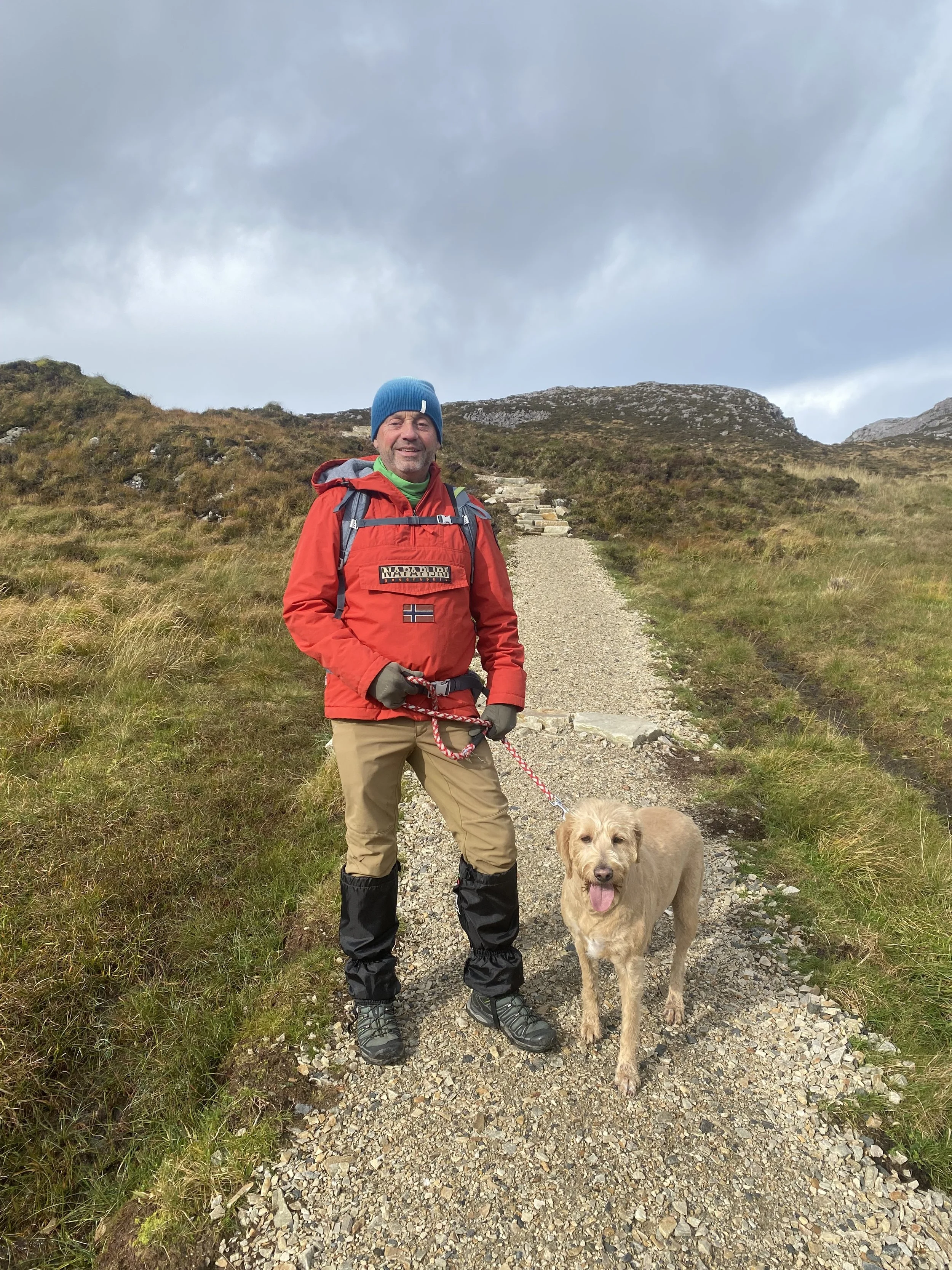 A man in outdoor hiking gear standing on a gravel trail with a golden retriever dog during daytime outdoor hike. The man is smiling and wearing a blue beanie, red jacket, and tan pants. The dog is panting with its tongue out, and both are in a green, hilly landscape with cloudy sky.