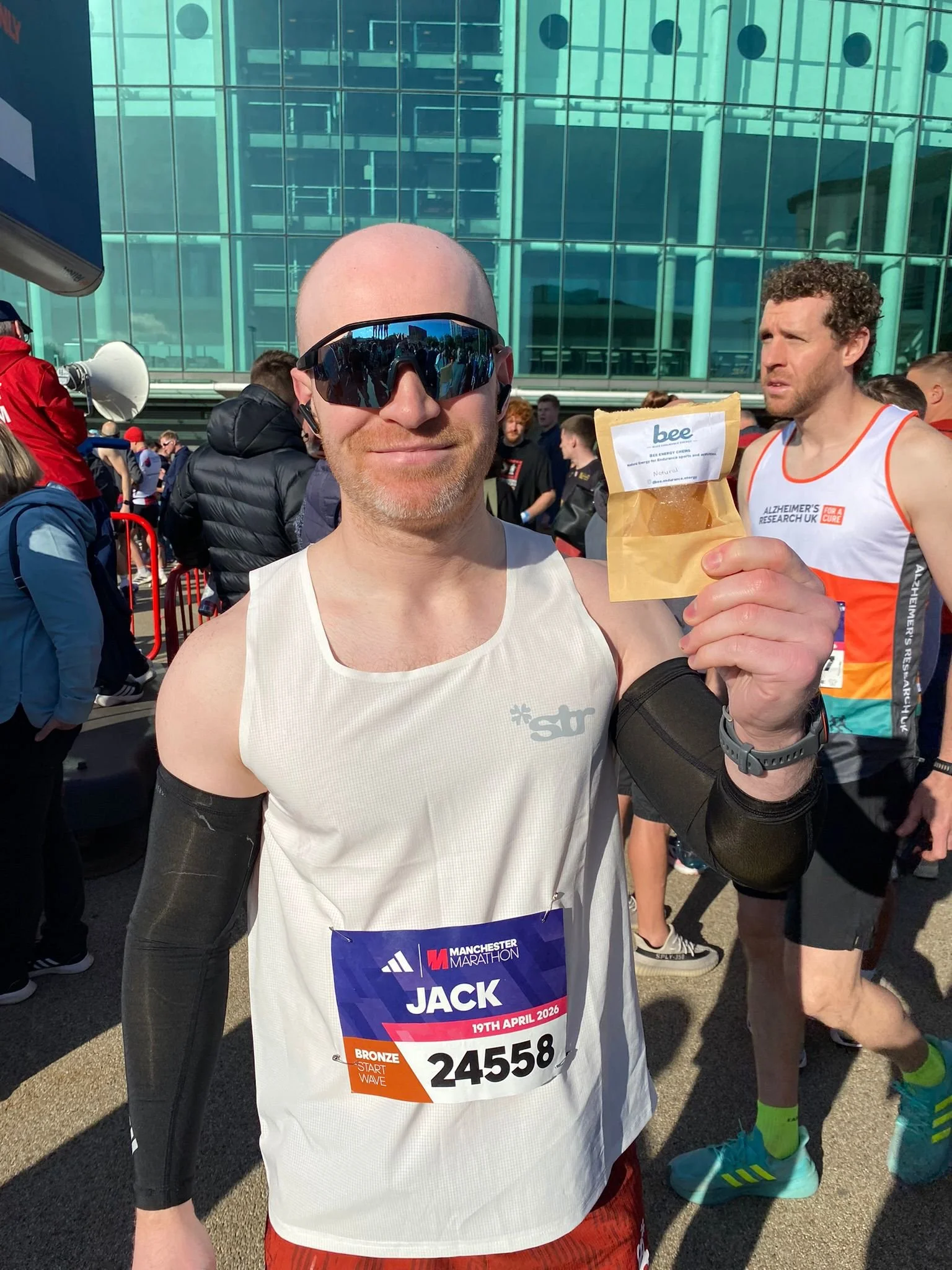 A man taking a selfie at the starting line of a marathon, wearing sunglasses and a race bib with his name 'Jack' and the date '19th April 2023', surrounded by other runners outside a modern glass building.