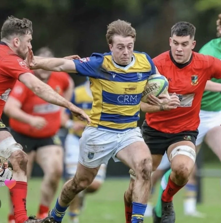 A rugby game featuring players running on the field, one in a blue and yellow jersey holding a rugby ball as he is tackled by players in red jerseys.