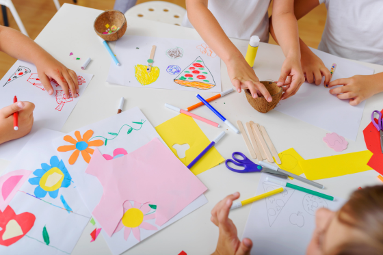 Children engaged in arts and crafts activities with paper, markers, scissors, and glue on a white table.