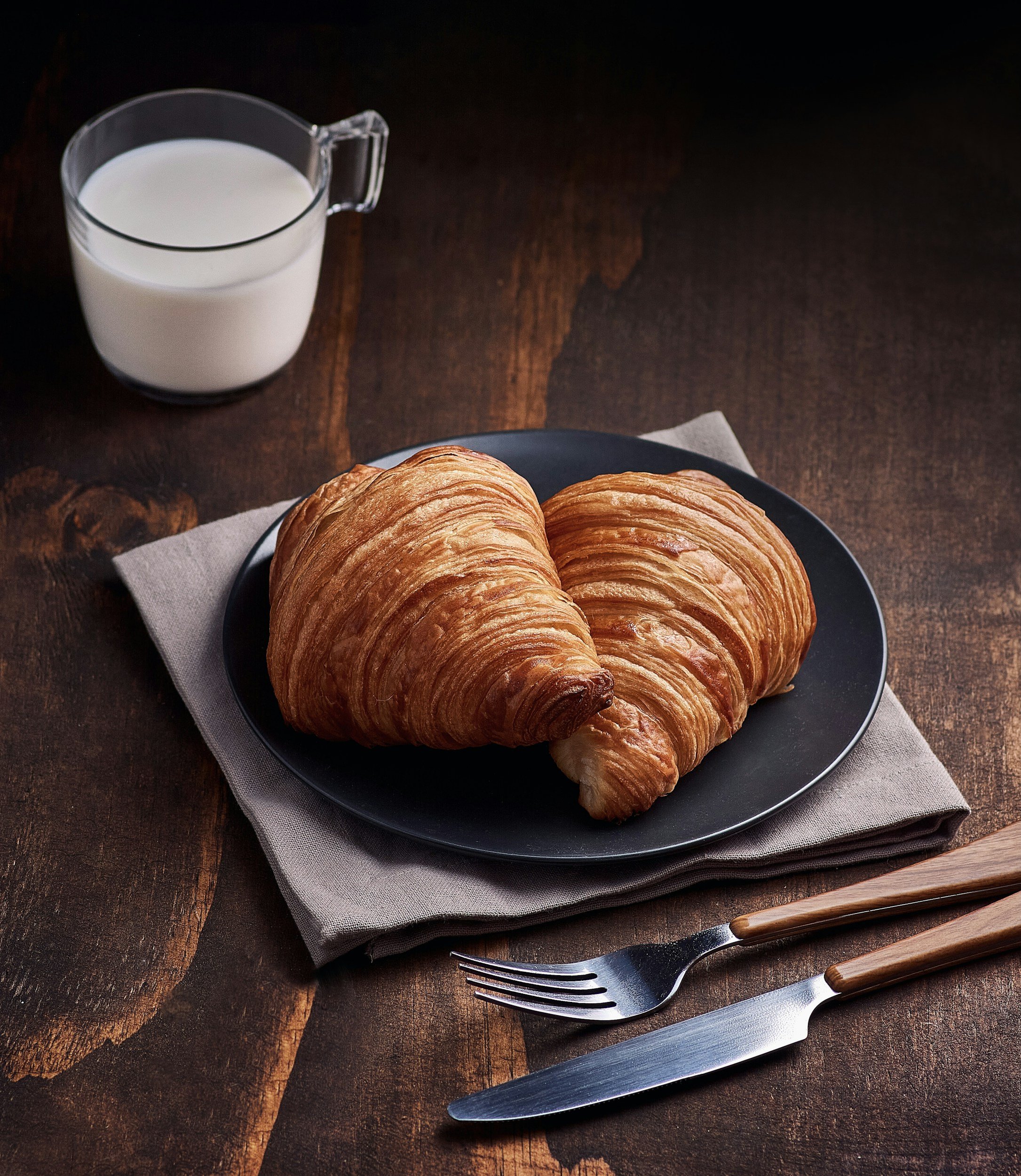 A black plate with two croissants on a gray napkin, a glass pitcher of milk in the background, and a fork and knife with wooden handles on a rustic wooden table.