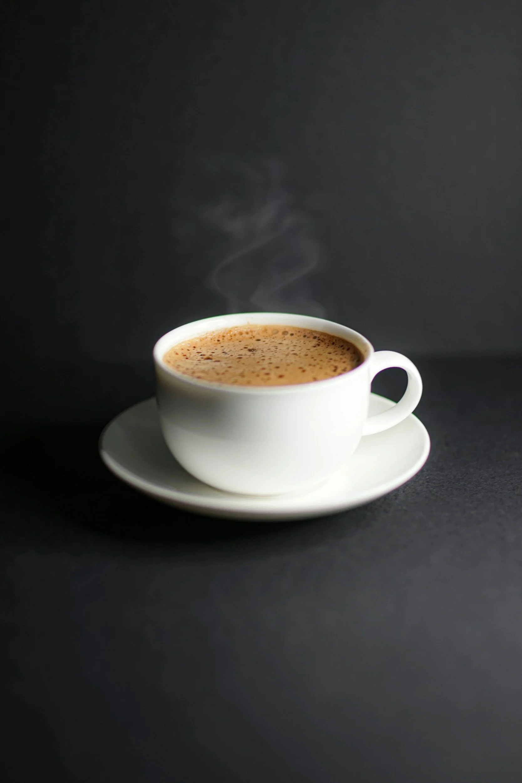 A white cup filled with hot coffee on a matching saucer, with steam rising from the surface, against a dark background.