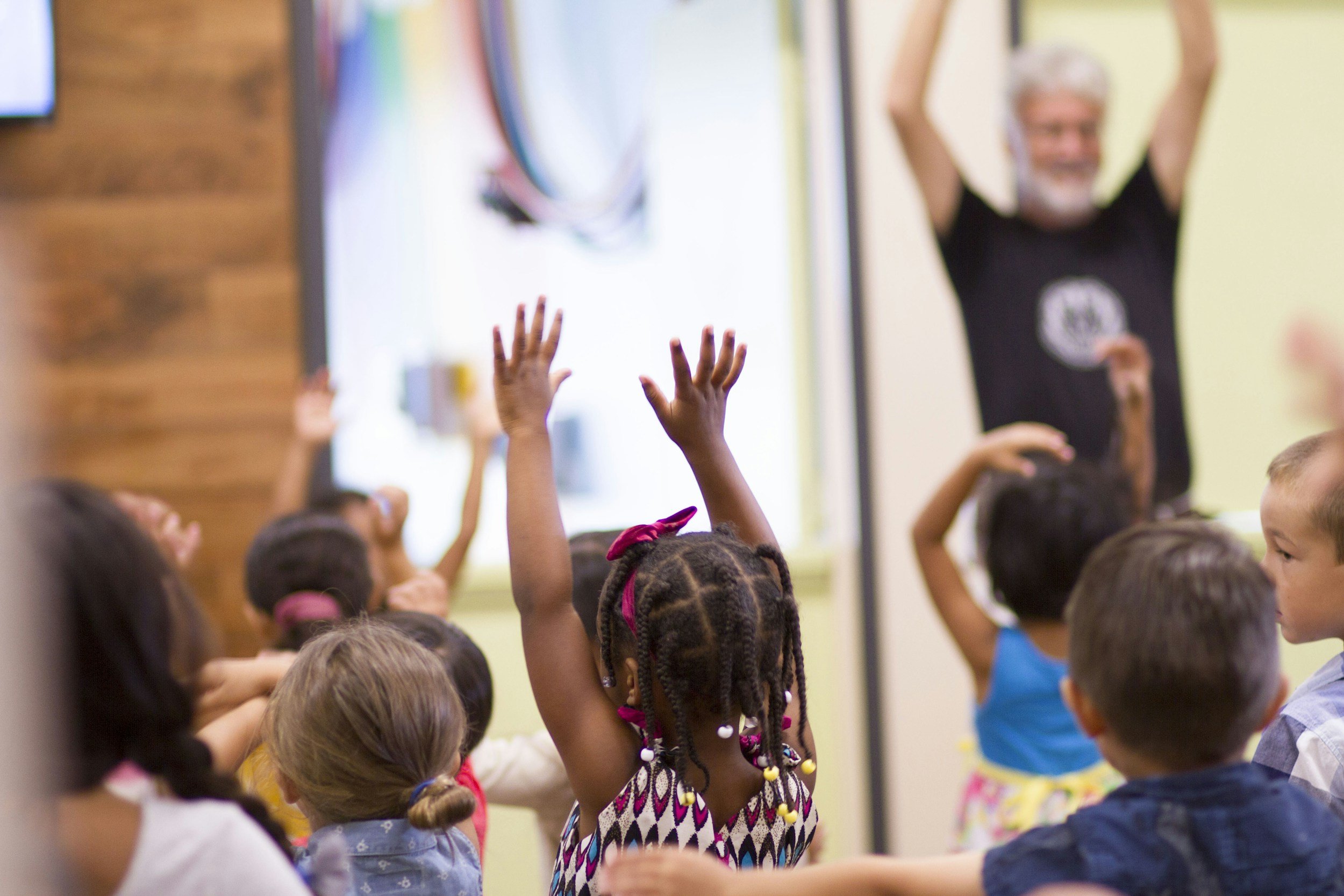 A classroom filled with young children raising their hands, with a teacher standing in front of a whiteboard.