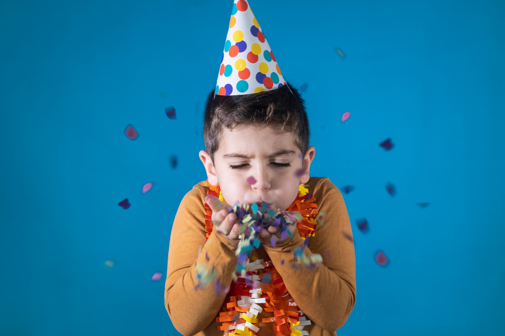 Boy wearing a party hat and colorful garland blowing confetti in celebration on blue background