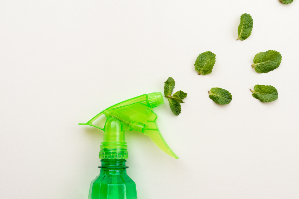 Green spray bottle with mint leaves scattered on a white surface.