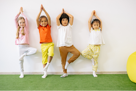 Four children practicing yoga in a line against a white wall, each standing on one leg with hands together above their heads.
