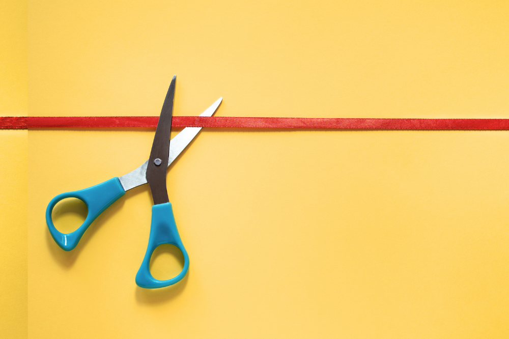 Pair of scissors cutting a red ribbon on a yellow background.