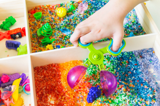 A hand using a green scoop to play with colored glitter and sand in a sensory bin, with small toys in the background.