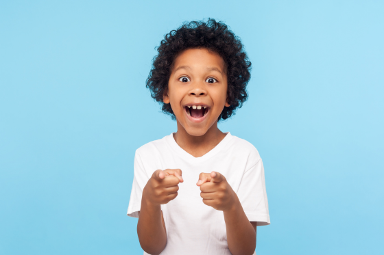 Joyful young boy with curly hair smiling and pointing at the camera against a blue background.