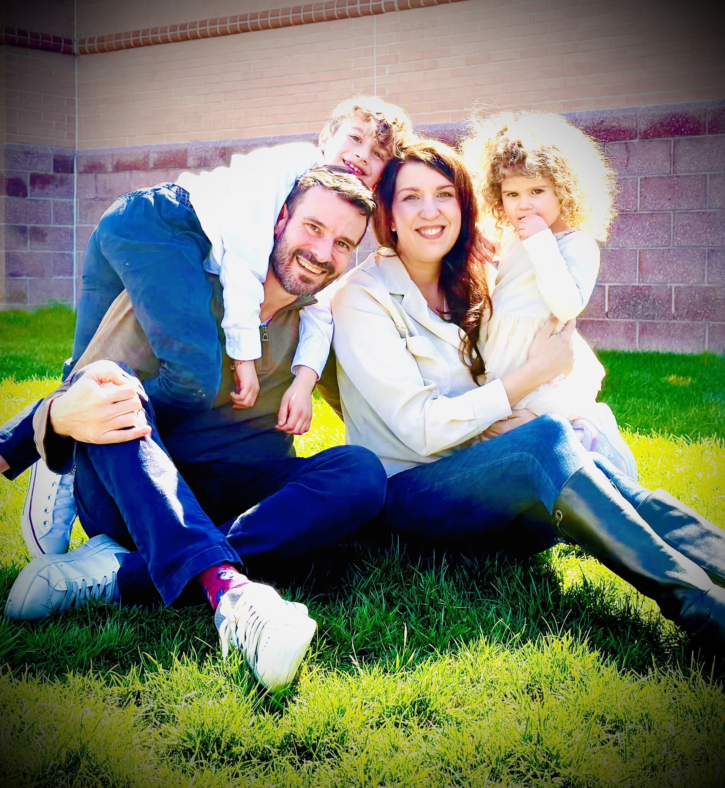 Family of four sitting on grass, smiling, with a brick wall behind them. The family includes two parents and two children, a boy and a girl, enjoying a sunny day.