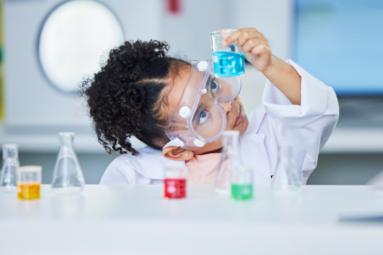 Young girl in a lab coat and safety goggles examining a beaker of blue liquid in a science lab.