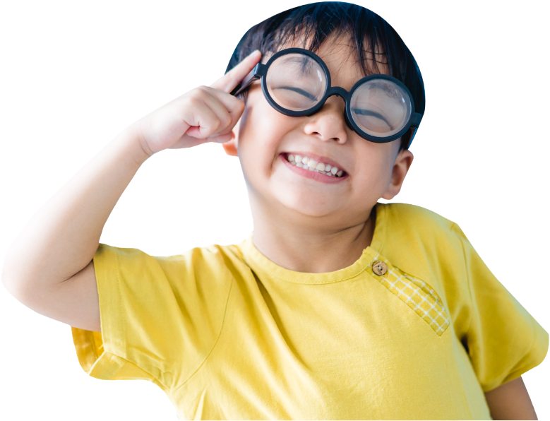 A young boy wearing large round glasses and a yellow shirt, smiling and pointing to his temple.