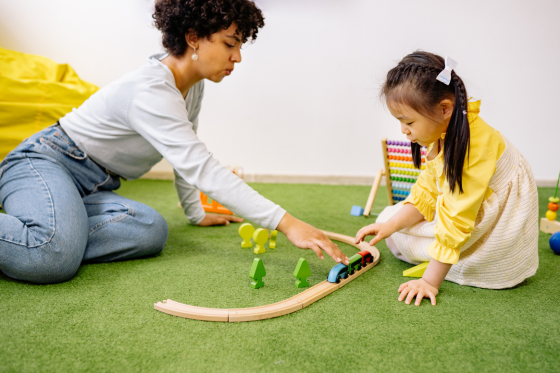 Adult and young girl playing with a wooden train set on green carpet.
