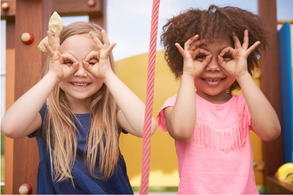 Two young girls playing on a playground, making funny faces by holding their hands around their eyes, smiling, with a wooden play structure and yellow background.
