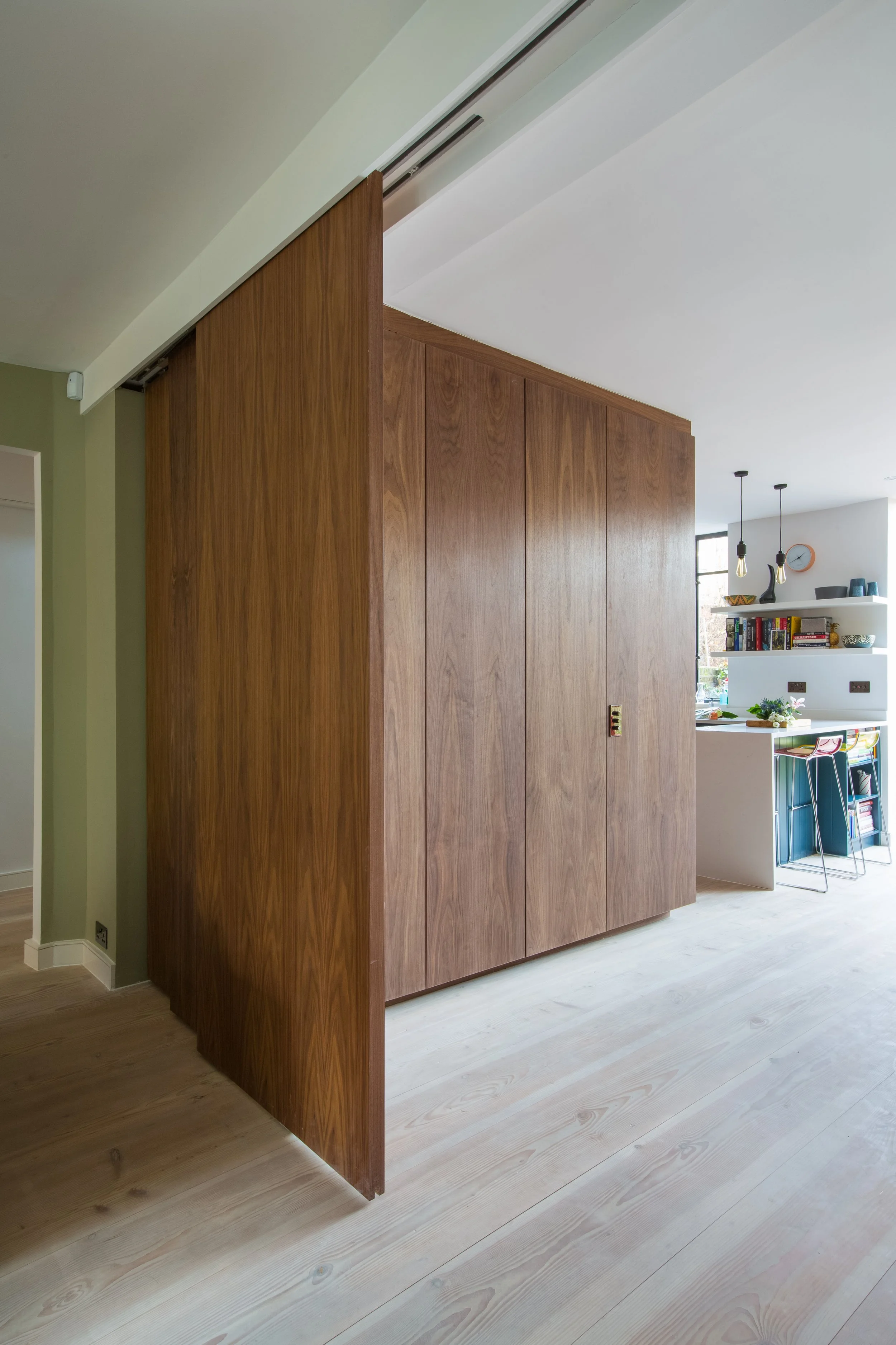 Interior view of a kitchen with a large wooden sliding door dividing the space, light hardwood flooring, and a white wall with shelves holding books and decorative items in the background.