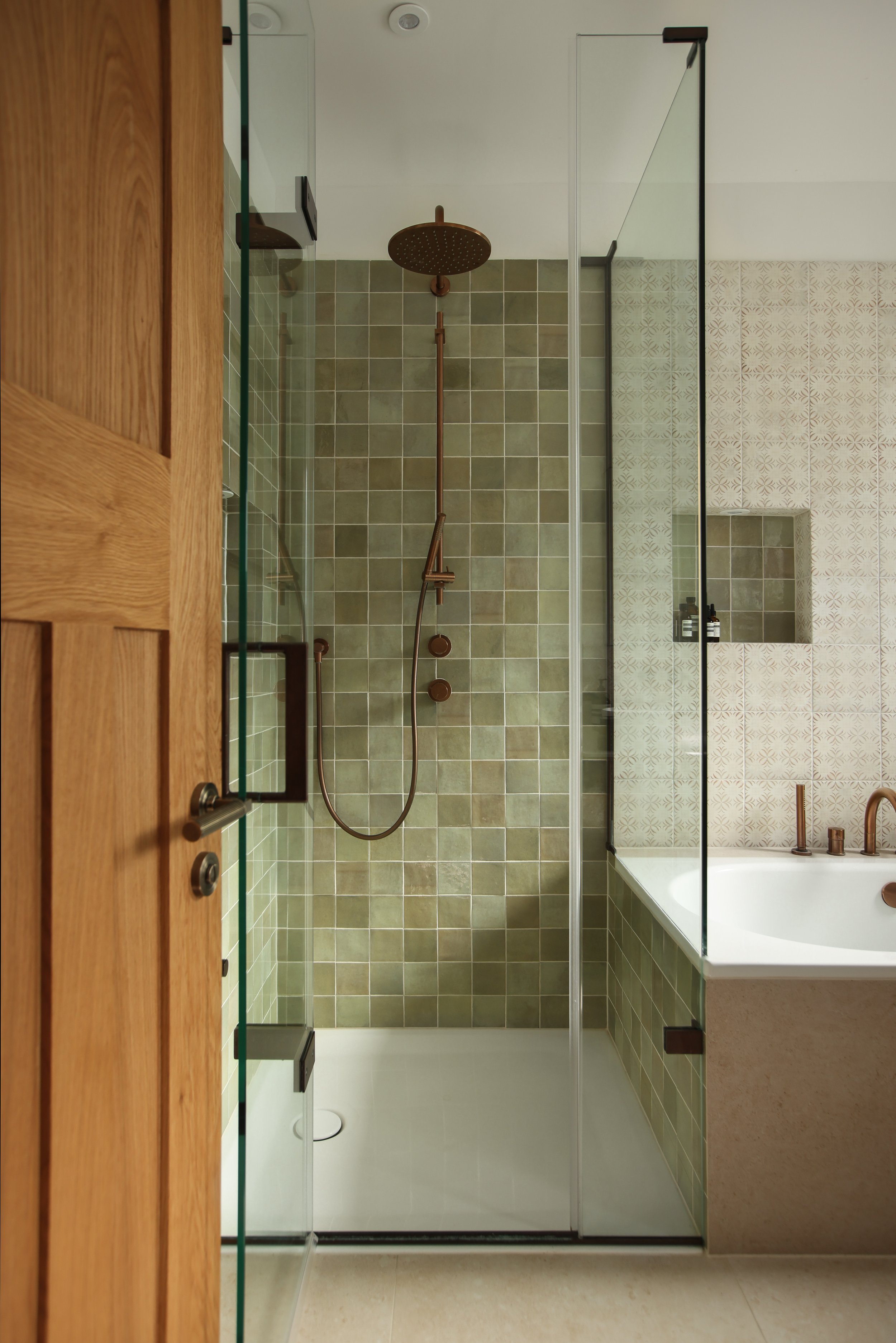 A modern walk-in shower with green tile, a rainfall shower head, a handheld shower, and a small shelf. Adjacent to the shower is a bathtub with a built-in shelf and two bottles. The bathroom has a large mirror, a wooden door frame on the left, and a small window on the right.