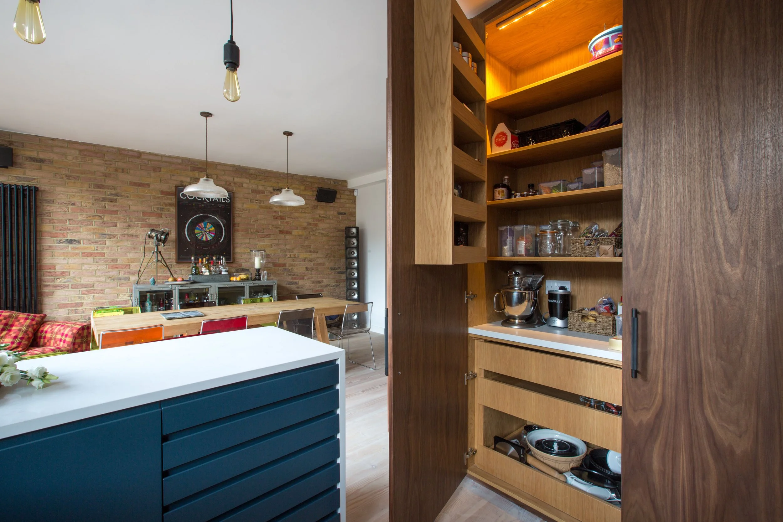 Open wooden pantry door revealing kitchen shelves with jars and containers, part of a modern kitchen with a brick wall, hanging pendant lights, and a dining table.