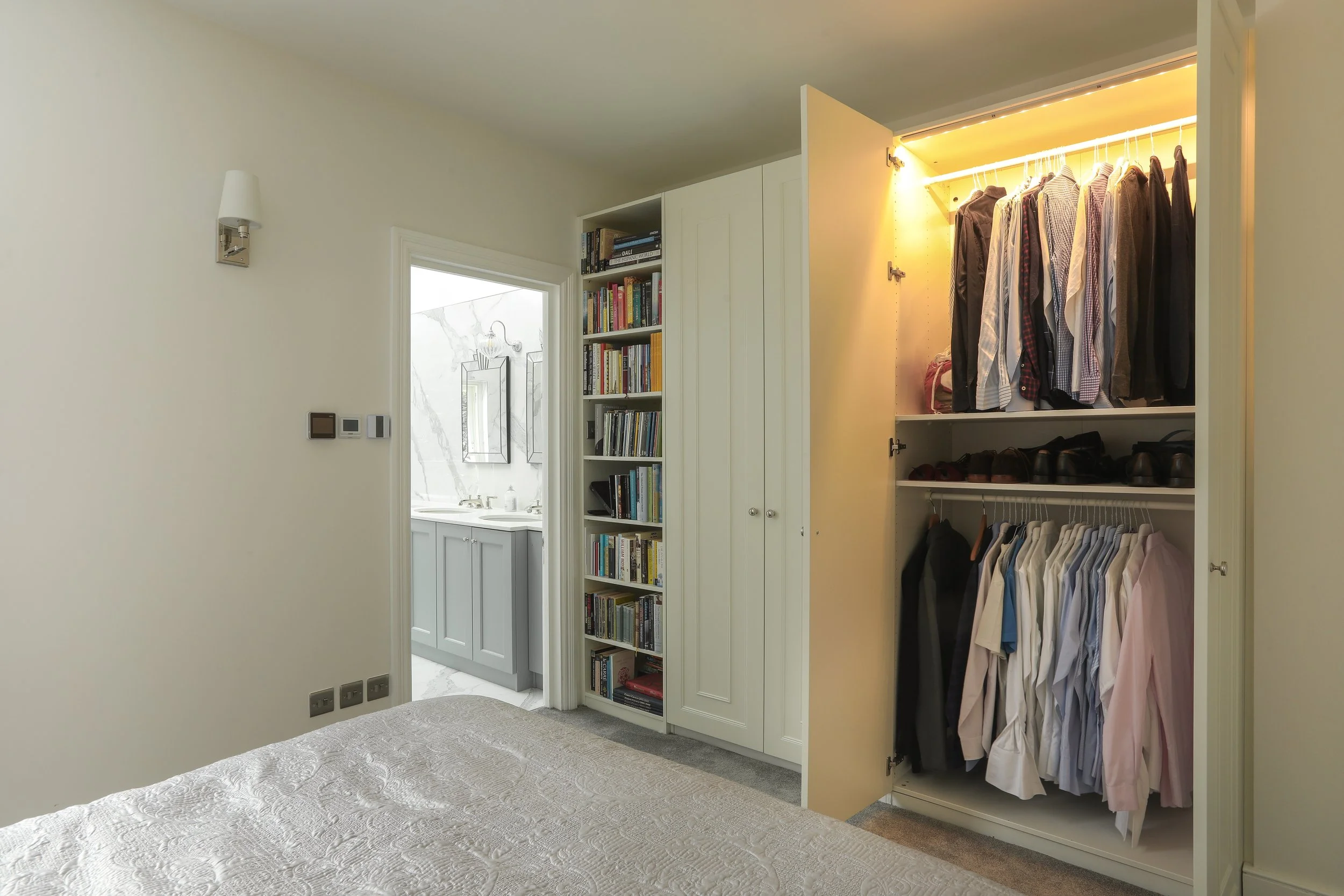 Bedroom with an open closet containing hanging shirts and shoes, a built-in bookshelf, and a view into a bathroom with a marble wall and a mirror.