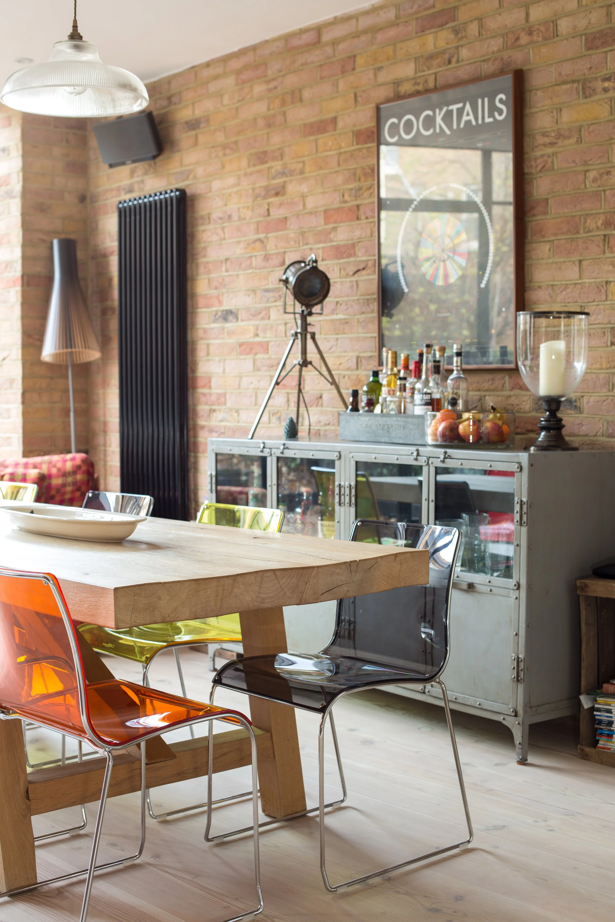Interior of a modern dining room with a large wooden table, colorful translucent chairs, brick walls, a bar cart with bottles and fruit, a painting with the word 'COCKTAILS,' and decorative lighting fixtures.
