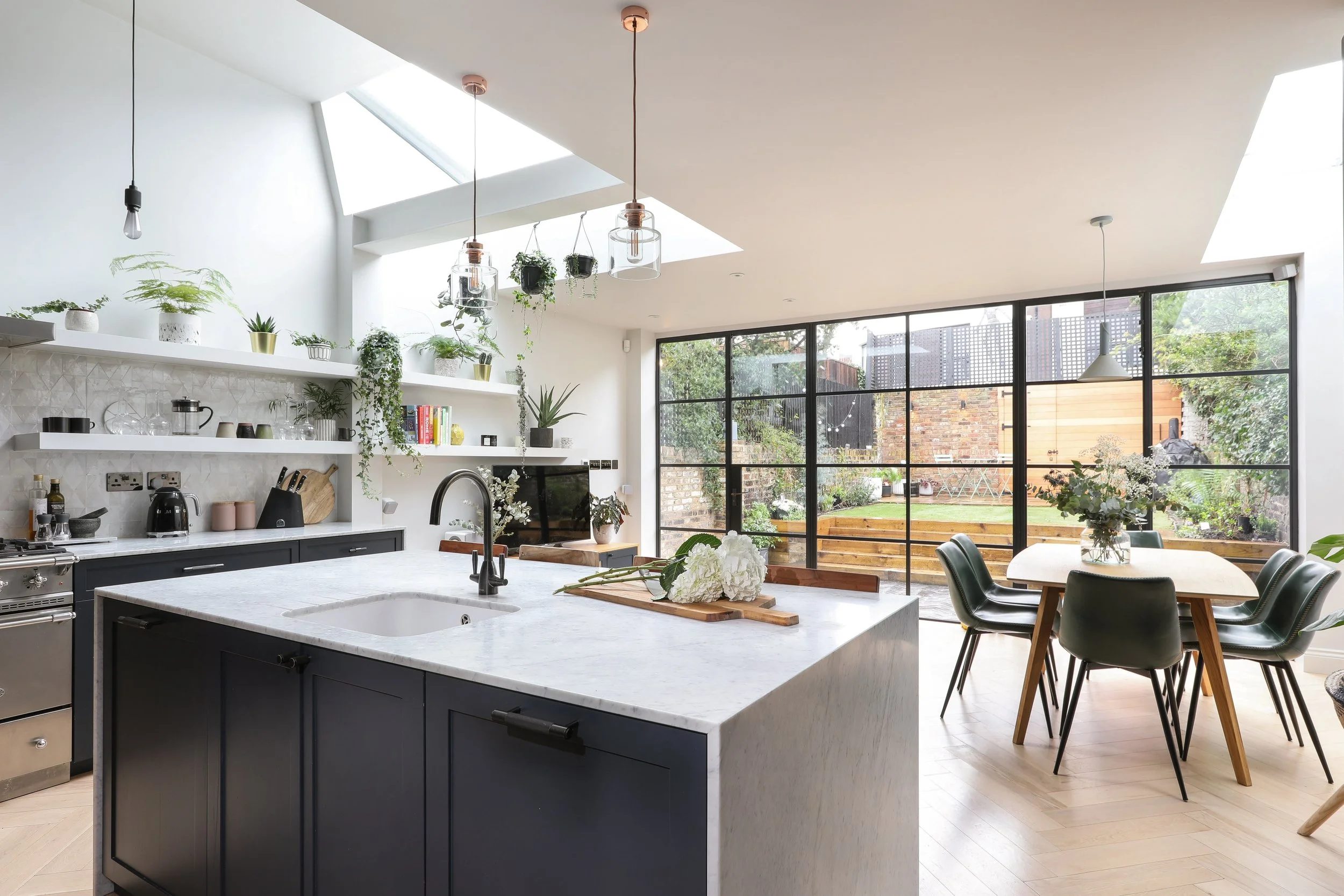 Modern kitchen with white walls, black cabinets, a white marble island, and open shelves with plants and kitchenware. Large glass sliding doors lead to a patio with outdoor seating and a backyard.