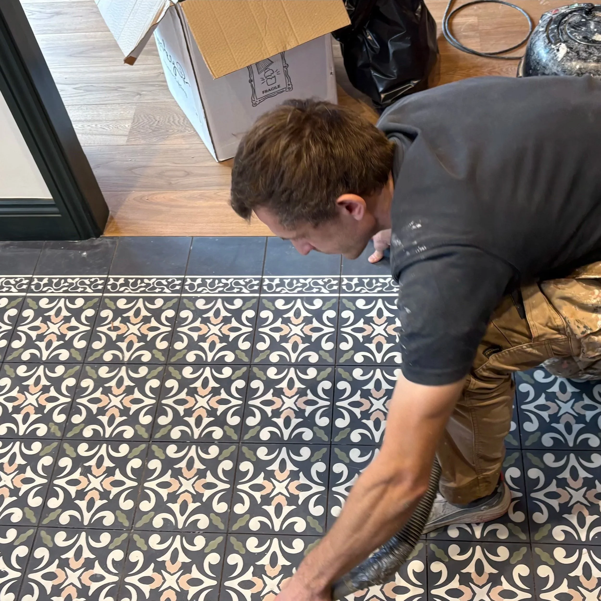 A person working on a tiled floor near a threshold, surrounded by boxes and trash bags in a room with hardwood and tiled flooring. Tiles are moroccon cement tiles from the mosaic factory. 
