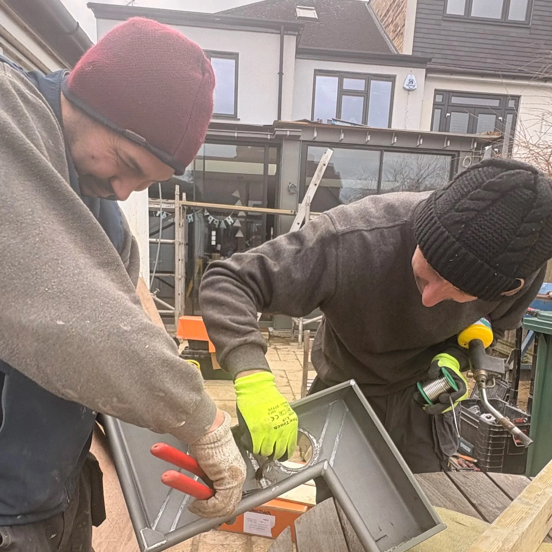 Two construction workers wearing safety gear working on a scaffolding outside a house. One is wearing a red beanie and glasses, and the other has a cap and safety glasses. Construction tools and equipment are visible around them.