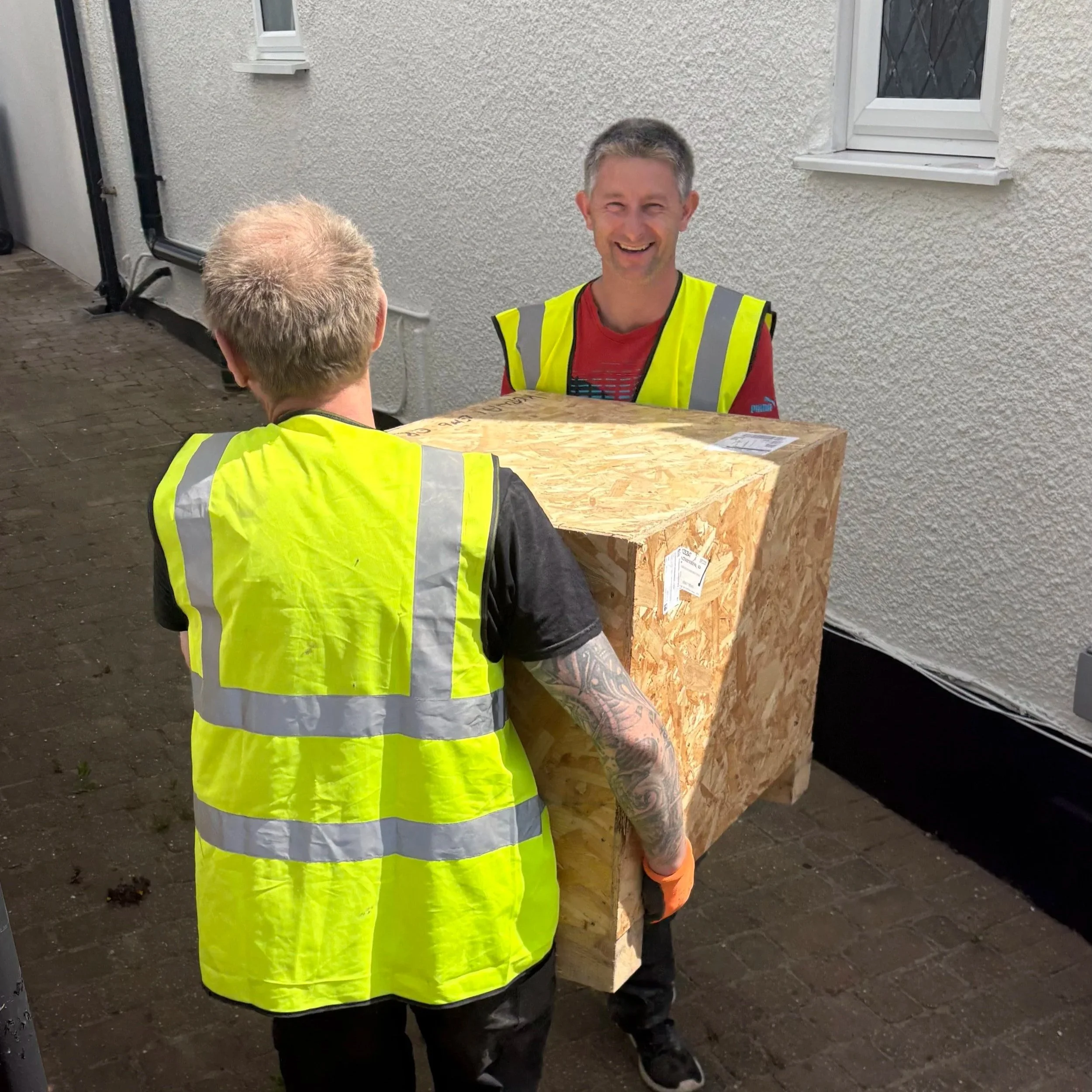 Two men, both wearing yellow safety vests, are standing outdoors near a light-colored textured wall with small windows. One man has gray hair and tattoos on his arm and is facing away. The other man has short, dark hair and is smiling, facing the cam