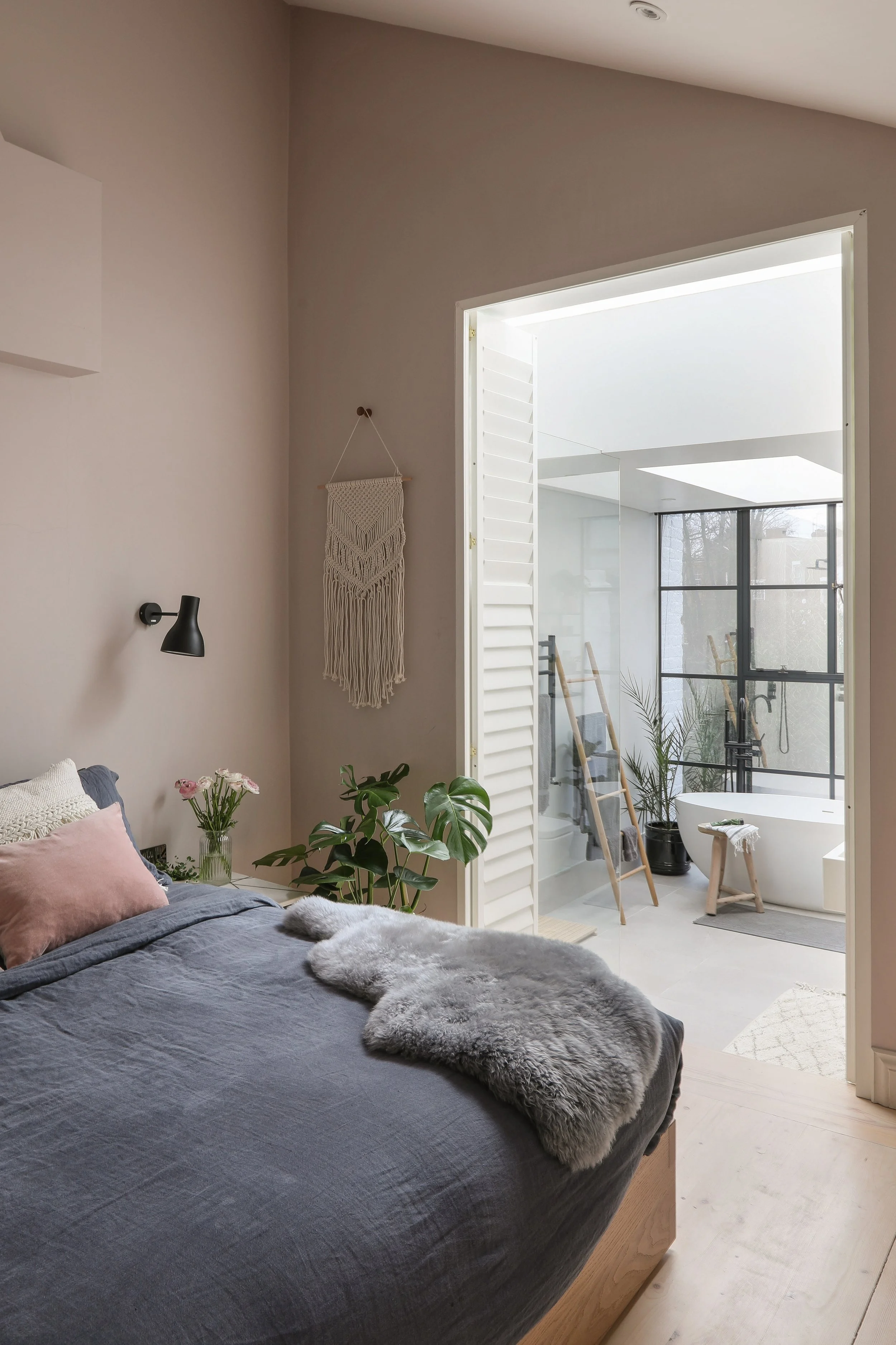 A cozy bedroom with a bed covered in a gray duvet, pink and white pillows, and a furry gray blanket. There are green plants on the bedside table and a wall hanging. A large window in the adjacent room allows natural light, revealing a modern, minimal