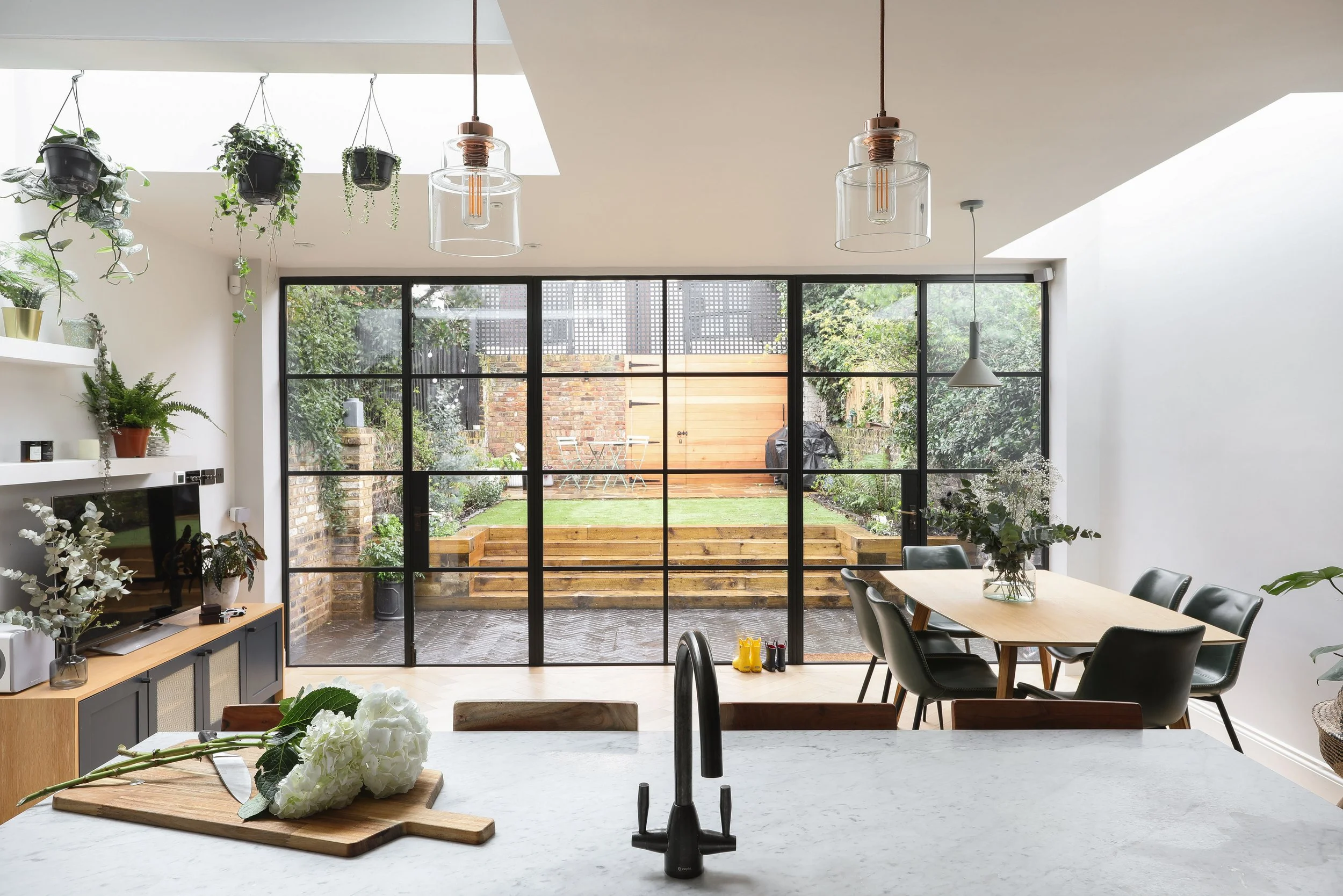A modern kitchen with a white marble countertop, black sink, and black faucet, decorated with white flowers on a cutting board, in front of a large glass sliding door leading to an outdoor patio and garden.