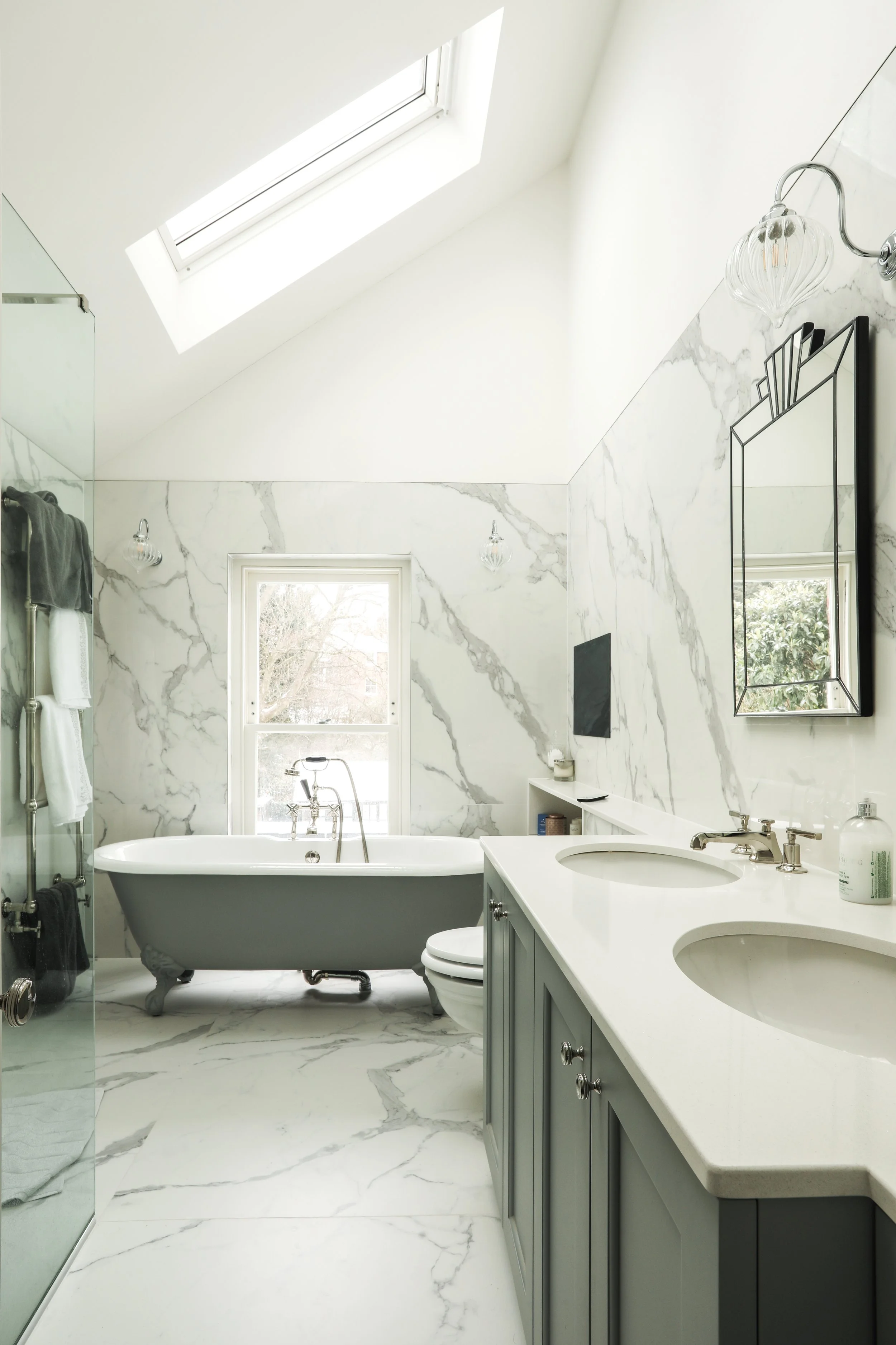 A luxurious bathroom with white marble walls and flooring, a freestanding bathtub with clawfoot style, a large window, a double sink vanity with gray cabinetry, a mirror, modern wall sconces, and a skylight ceiling with a view of the sky.