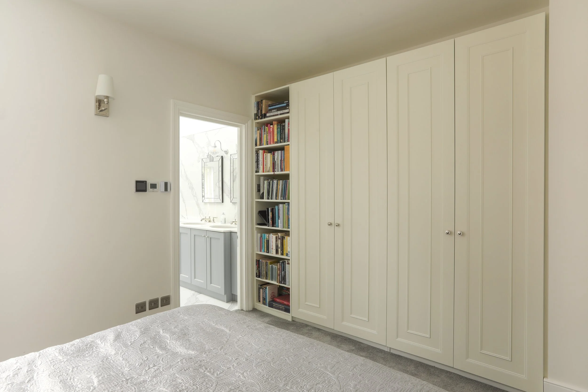 Interior view of a bedroom with white walls, a built-in white closet with paneled doors, a bookshelf filled with books, a doorway leading to a bathroom, and a bed with a textured white bedspread.