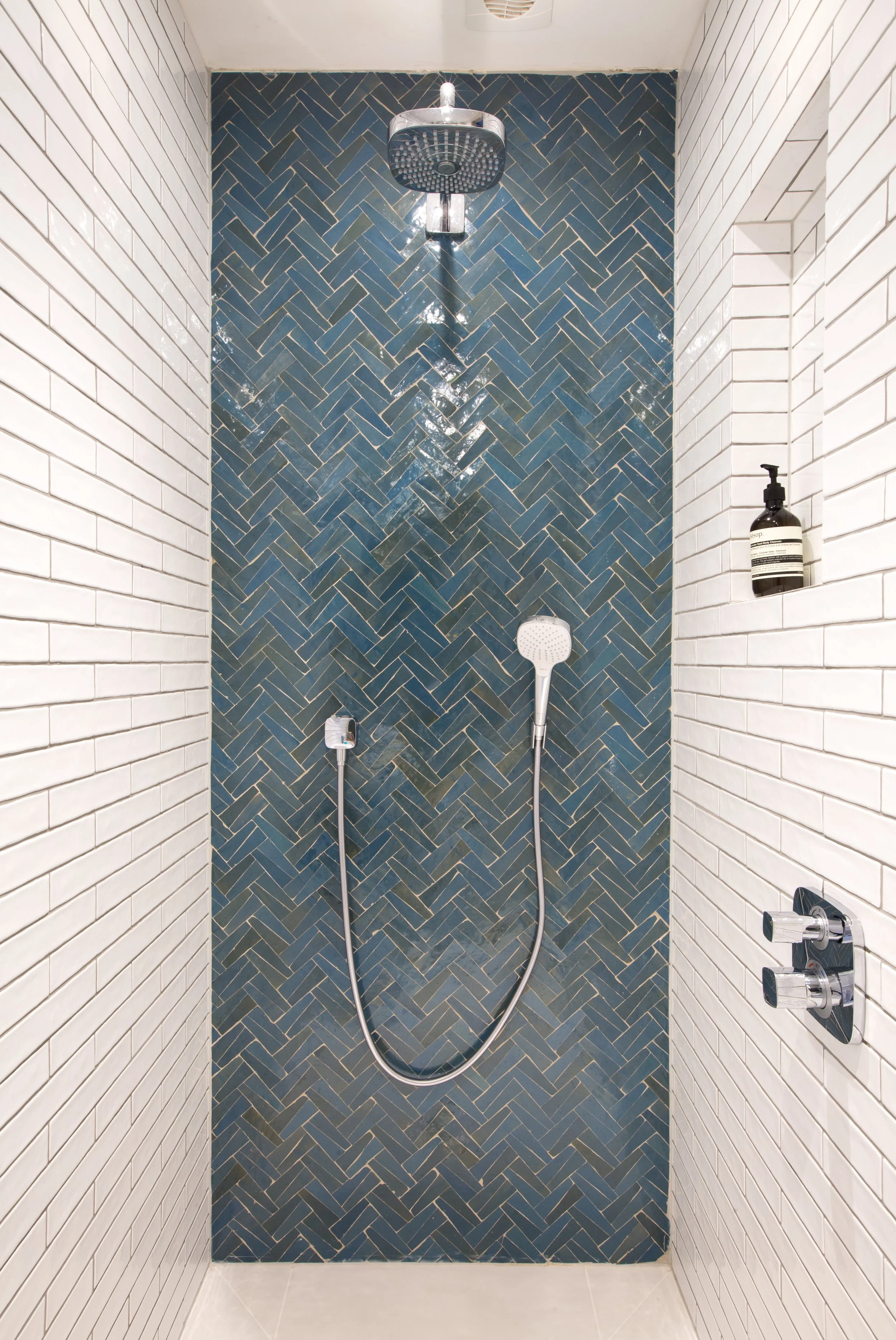 Shower area with blue herringbone tiles on the wall, white brick tiles on the side walls, a rain shower head, a handheld shower, and a bottle of shower product on a niche.