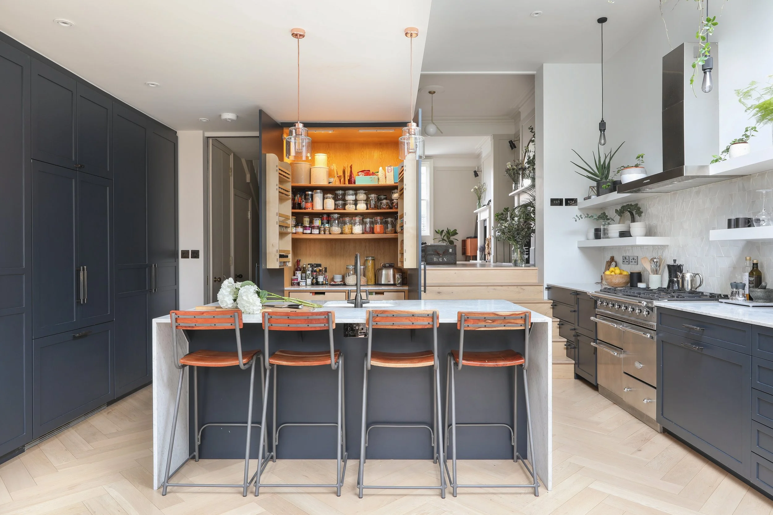 Modern kitchen with navy cabinets, a large island with a white marble top, and open shelving with plants and dishes. Wooden staircase leads to a pantry with jars of spices and ingredients. Hanging pendant lights and a gas stove add to the stylish des