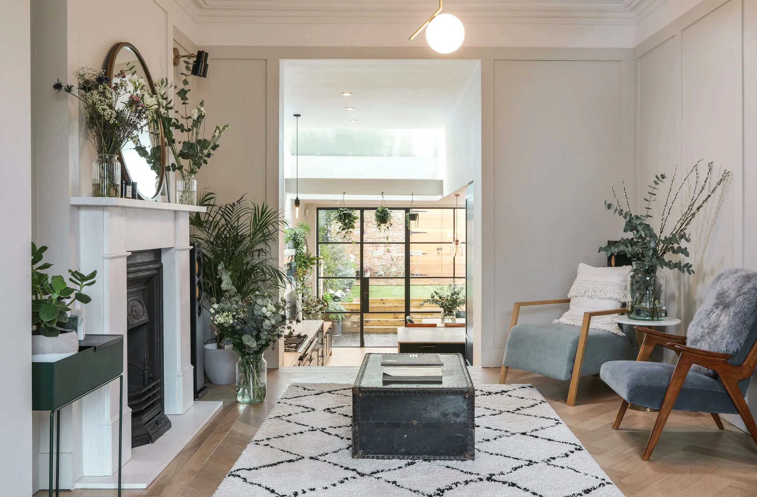 Interior view of a living room with a fireplace, plants, and a striped rug, leading to a dining area with hanging plants and a large window with a wooden privacy fence outside.