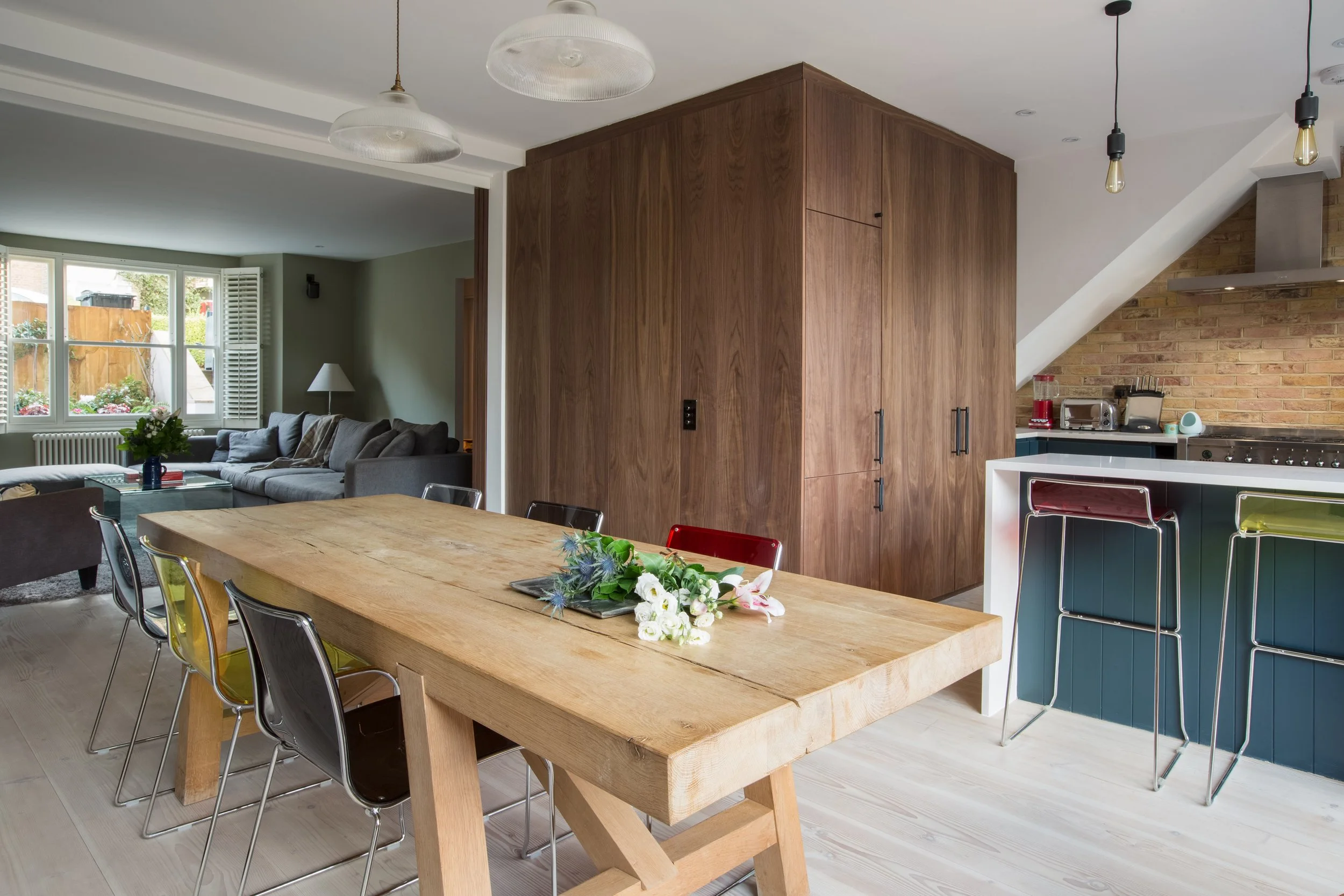 Open-concept kitchen and living room with wooden dining table, black and metal chairs, gray sofa, and large windows with white shutters.