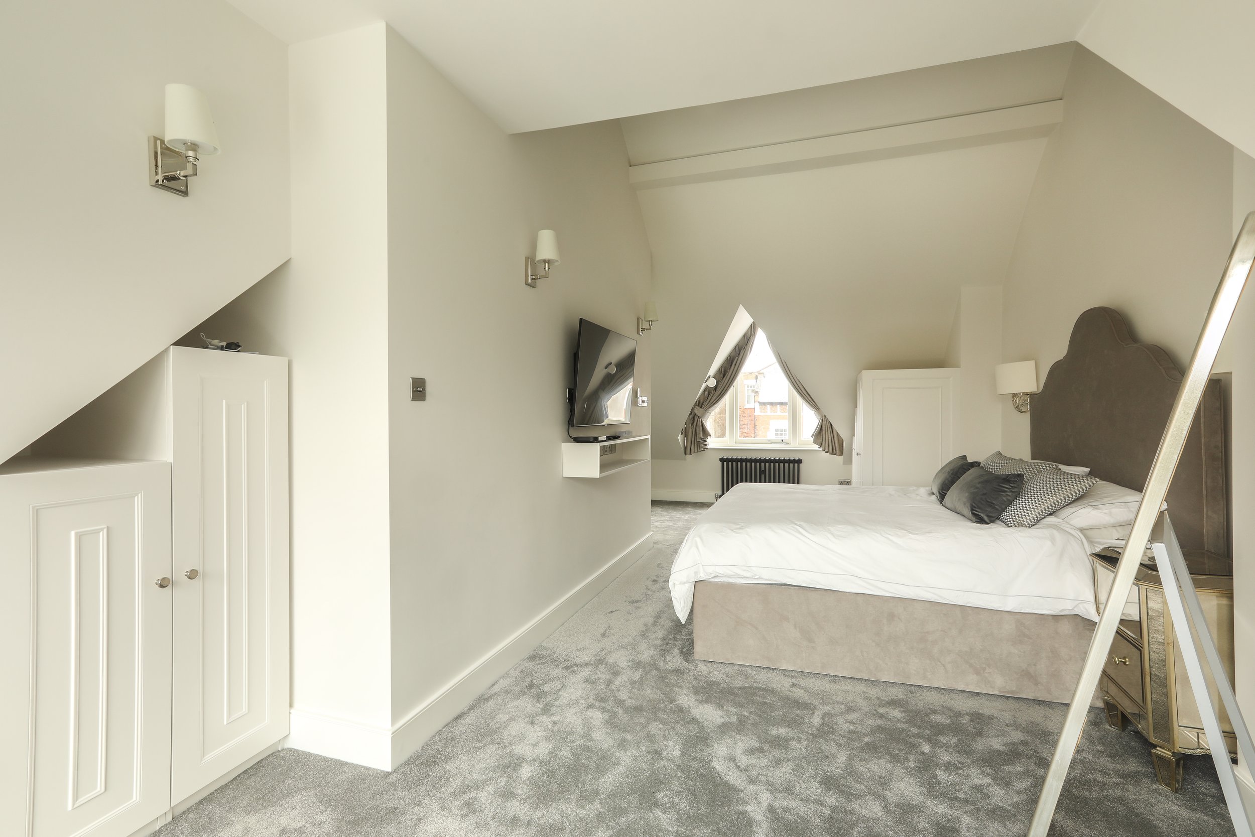 A bedroom with sloped ceilings, white walls, a large bed with grey headboard and pillows, a wall-mounted TV, a window with curtains, a small white cabinet, and a grey carpet.