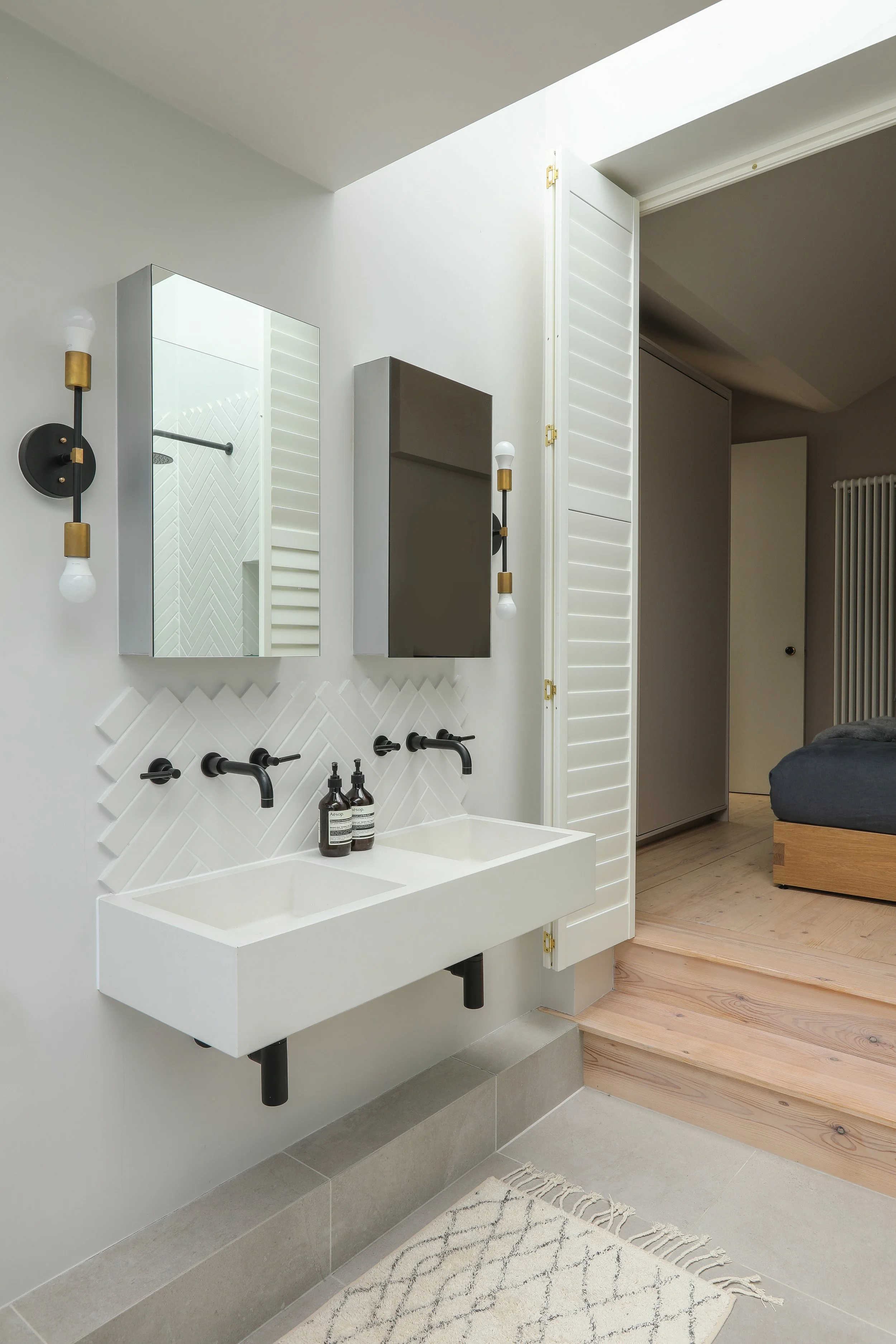 Modern bathroom with white rectangular sink, black fixtures, and two bottles of hand soap, with a mirror and a gray cabinet above the sink, wooden flooring, and a white wall with decorative tiling.