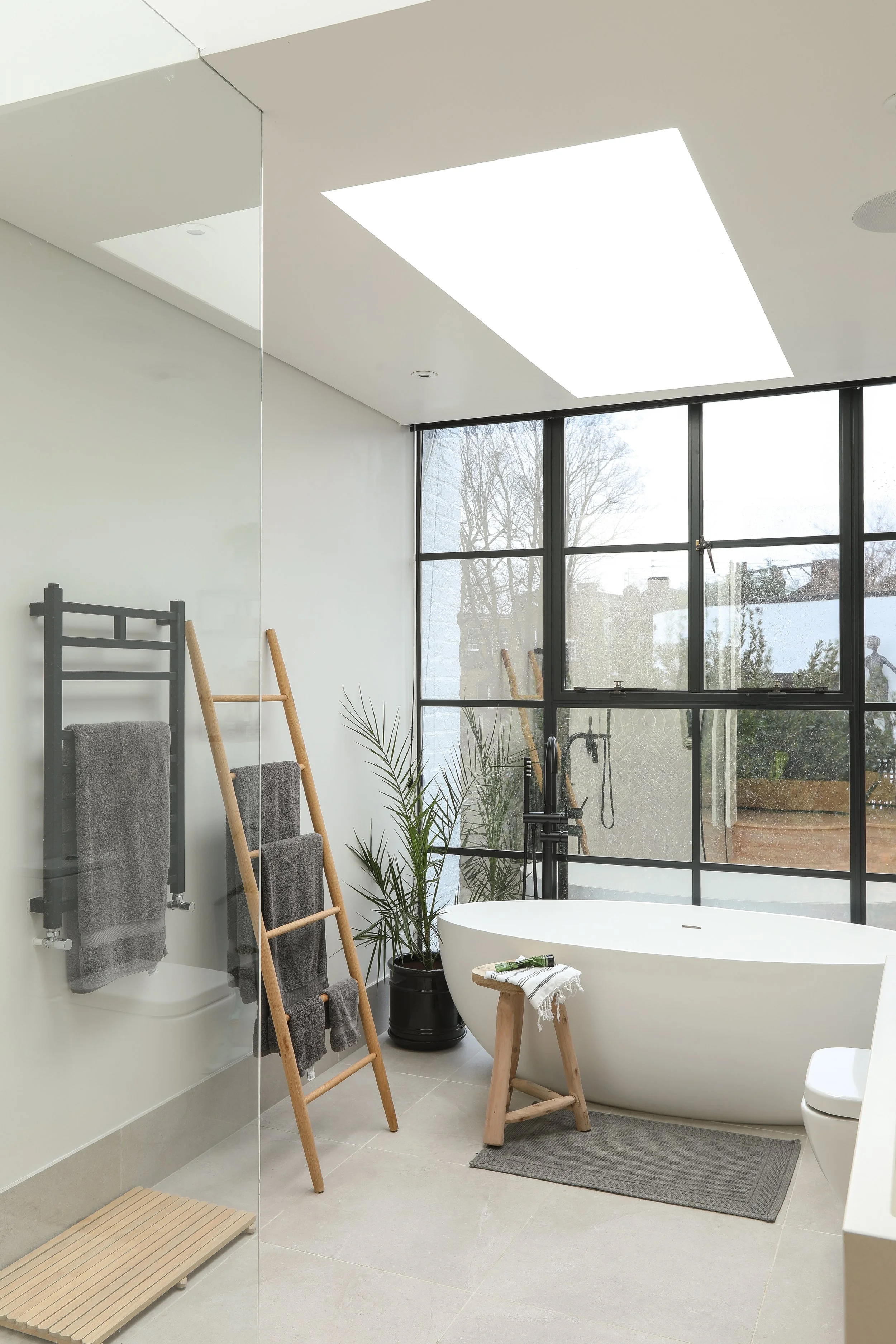 Modern bathroom with large bathtub, black window frames, and natural light. Includes wooden ladder, gray towels, potted plant, and small stool with towel and soap.
