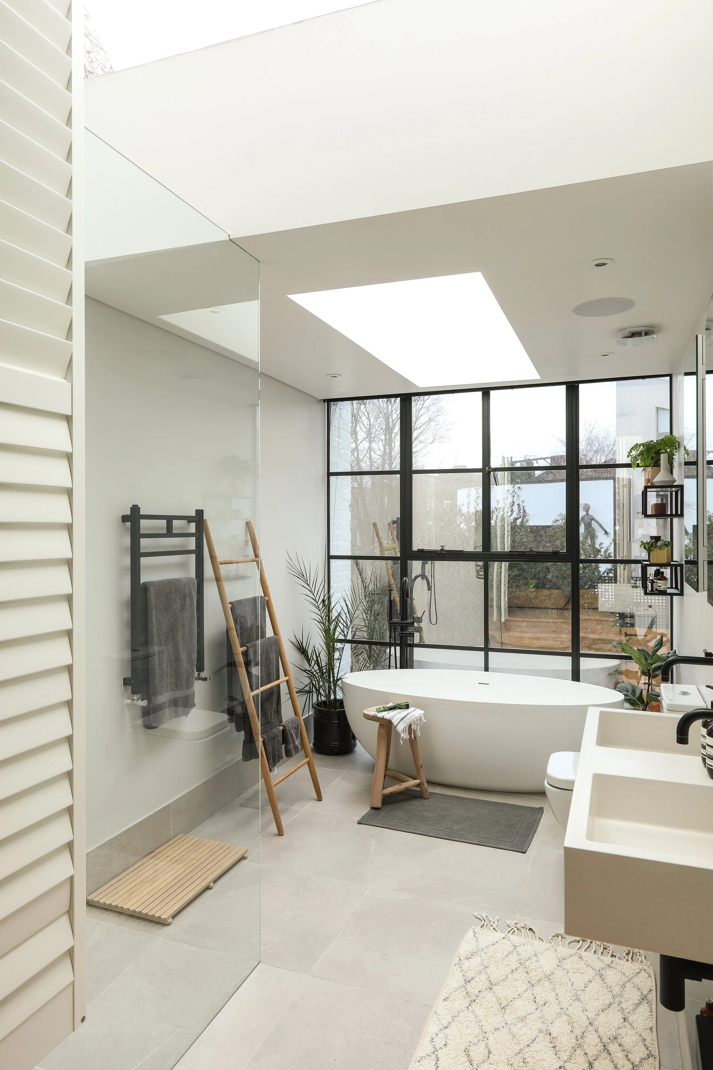 Modern bathroom with a bathtub, large window, and minimal decor, featuring plants, towels, and a stool.