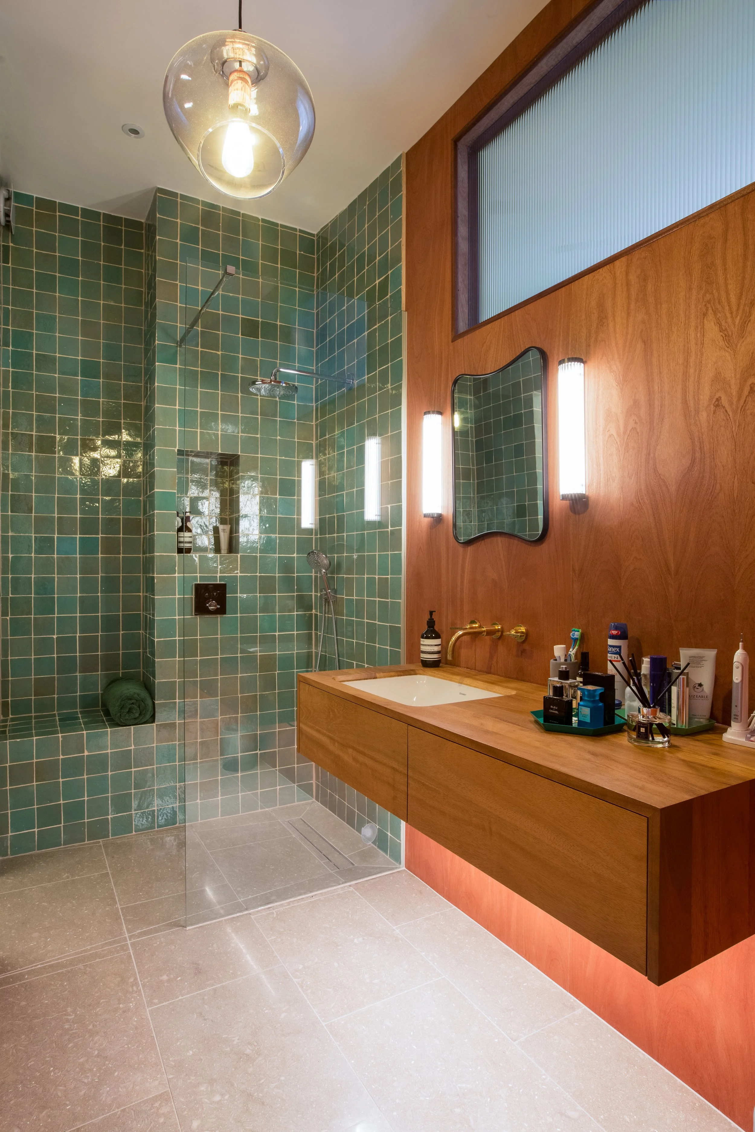 A modern bathroom with a green tiled shower area and a wooden vanity with a white sink. The vanity has a mirror, two vertical wall lights, and various toiletries on top. The ceiling features a glass light fixture, and there is a frosted window above 