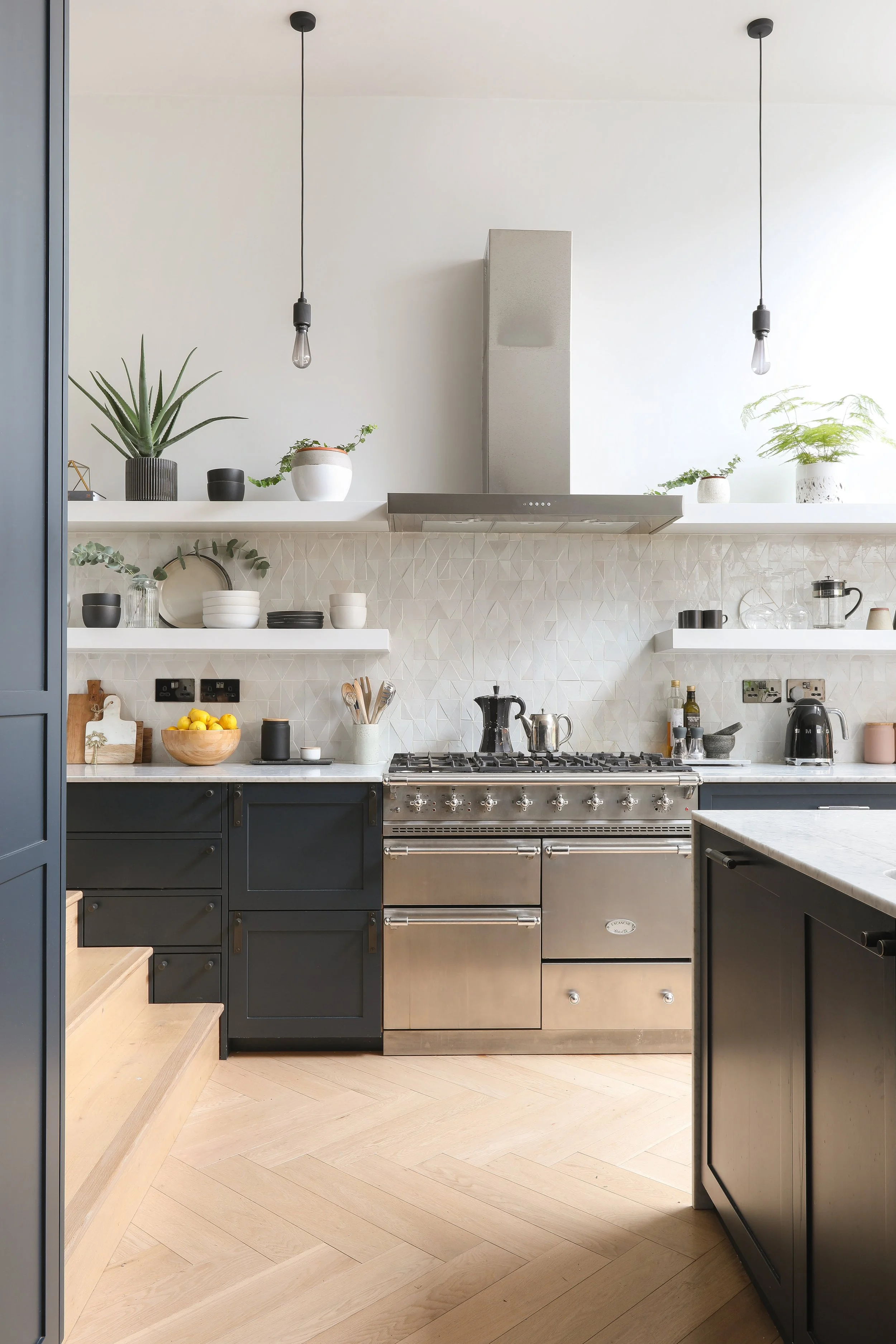 Modern kitchen with dark lower cabinets, open shelving, stainless steel oven, and light wood flooring. White walls with plants and two hanging light fixtures.