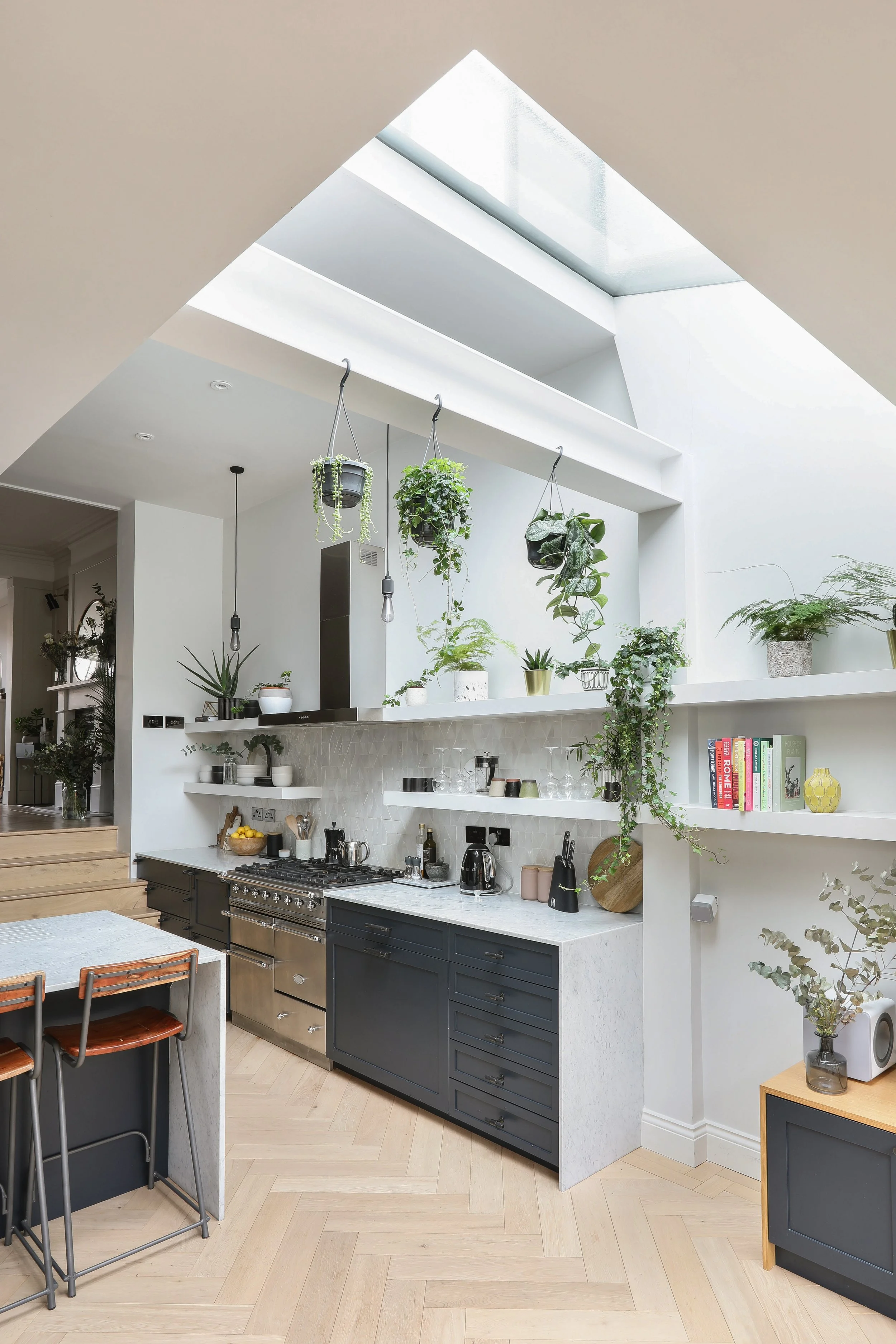 A modern kitchen with a white upper wall and ceiling. Hanging plants are above the counter. The kitchen features black lower cabinets, a stainless steel oven, and various kitchen utensils. A small bookshelf with colorful books is on the right, with a