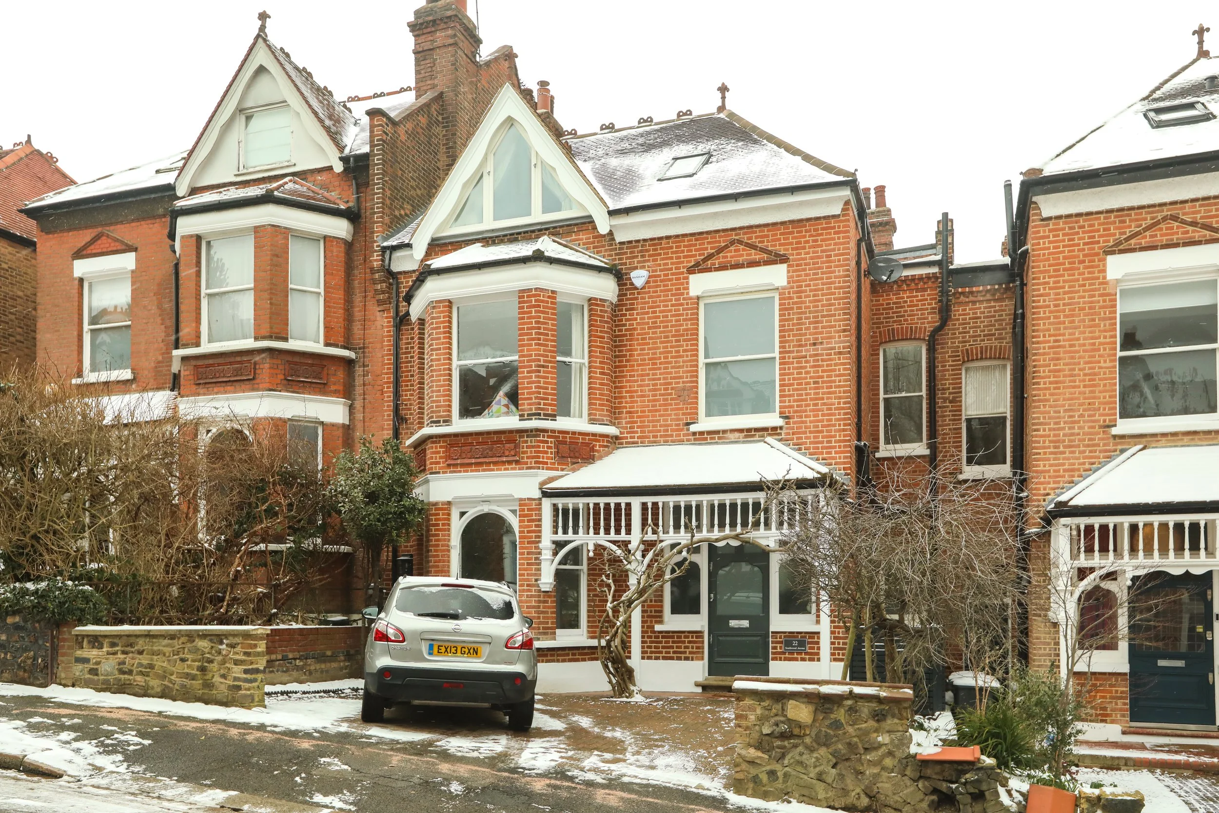A red brick Victorian-style house with a snow-covered roof, large bay windows, and white trim. There are cars parked in front, snow on the ground, and trees with no leaves.