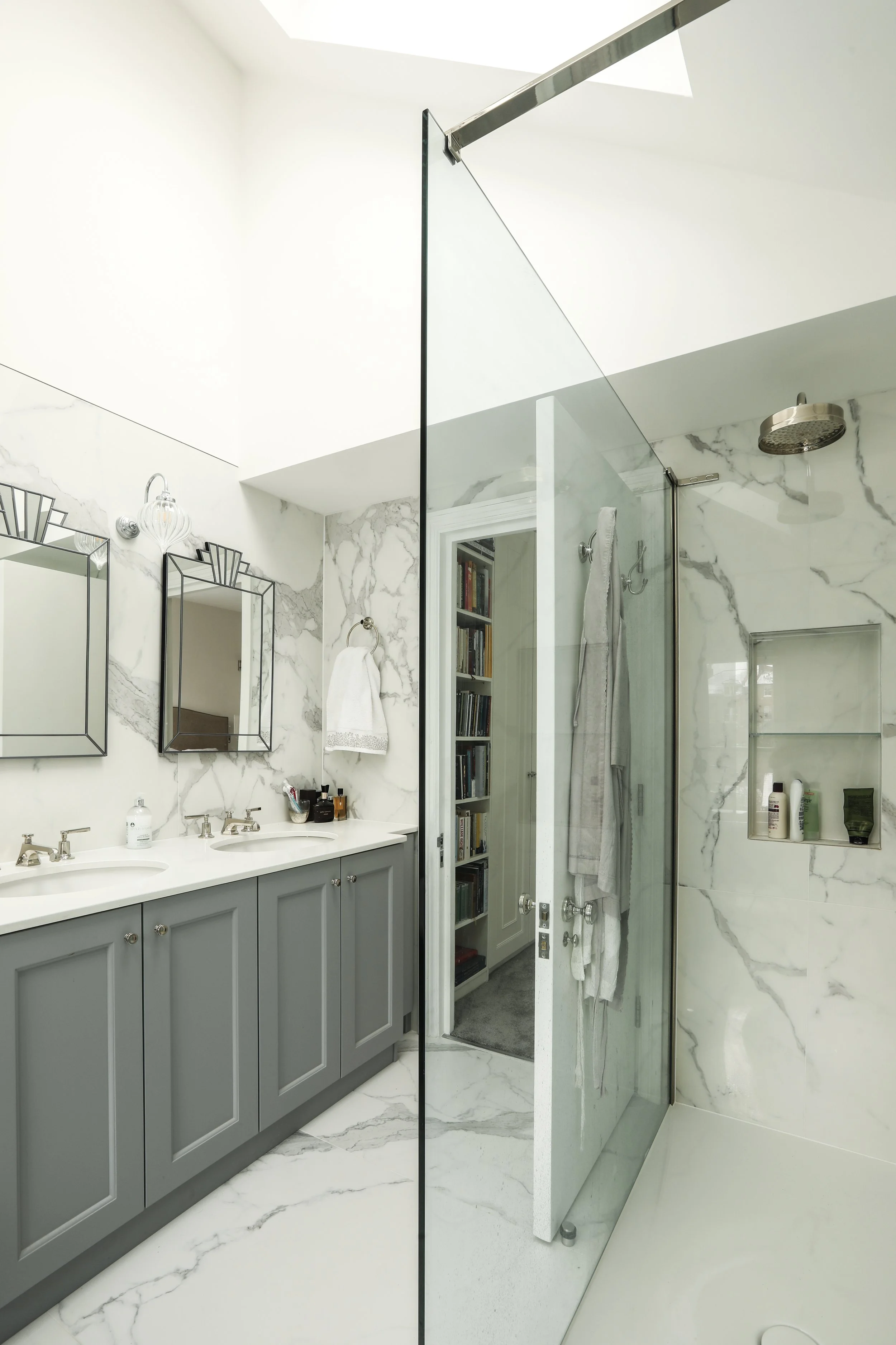 Modern bathroom with double sinks, marble countertops, gray cabinetry, mirrored medicine cabinets, a glass shower with marble walls, and minimalist fixtures.
