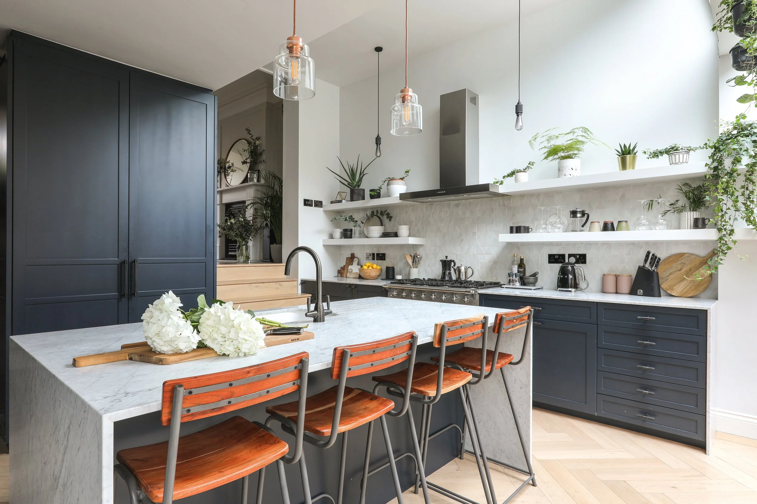 Modern kitchen with navy blue cabinets, marble island, and open shelves filled with plants, dishes, and glassware. Pendant and Edison bulbs hanging above, with a bouquet of white flowers on the island.