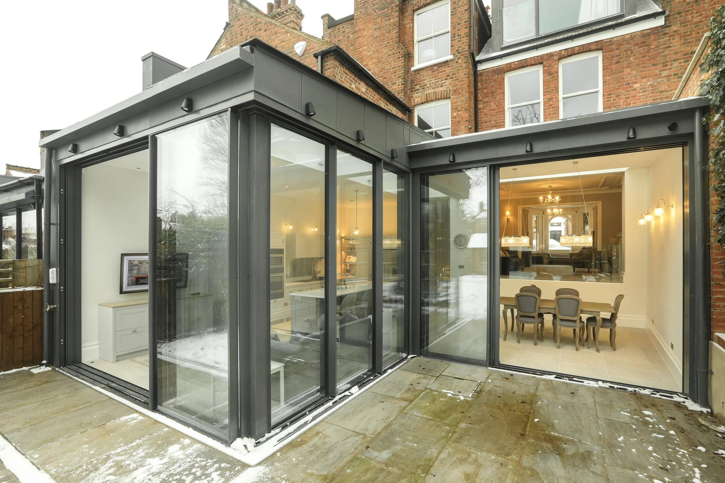 A modern glass extension attached to a traditional red brick house with large windows and a view of a dining room inside.